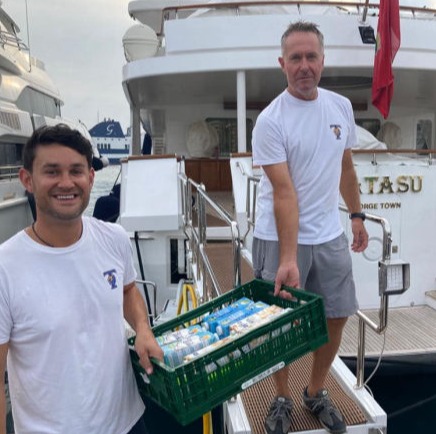 A volunteer from YGB stands in front of a yacht and next to bags of items donated by the yacht