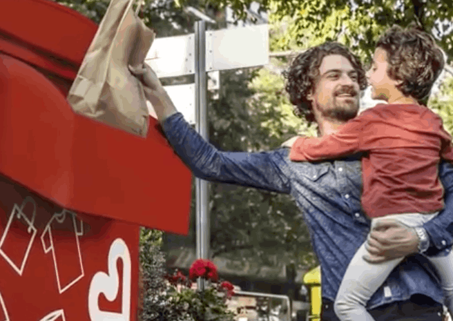 A man with a child in his arms drops items in a Caritas container